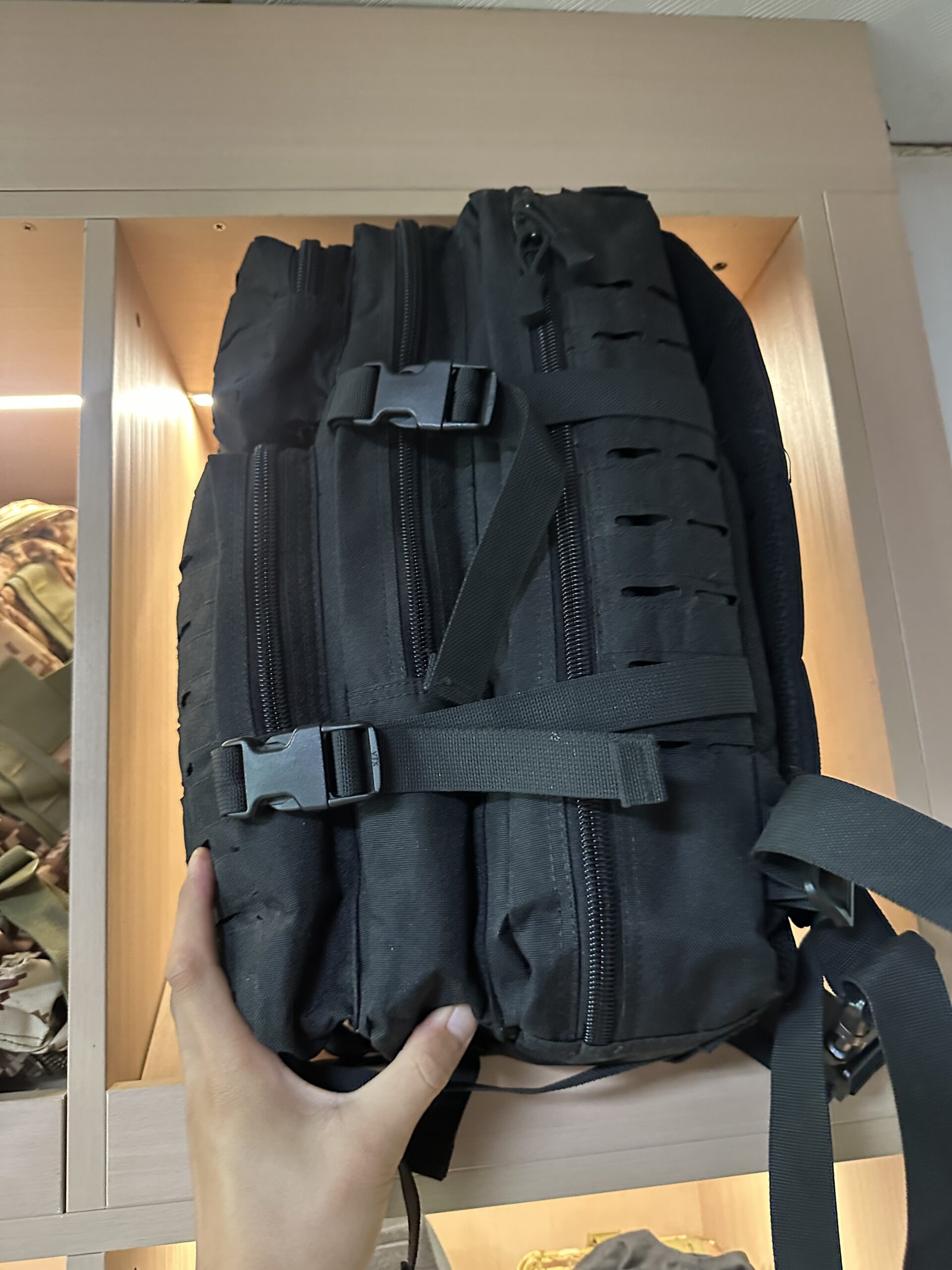 Close-up of a black tactical backpack folded compactly, showing multiple zippered pockets, MOLLE webbing, and buckle straps