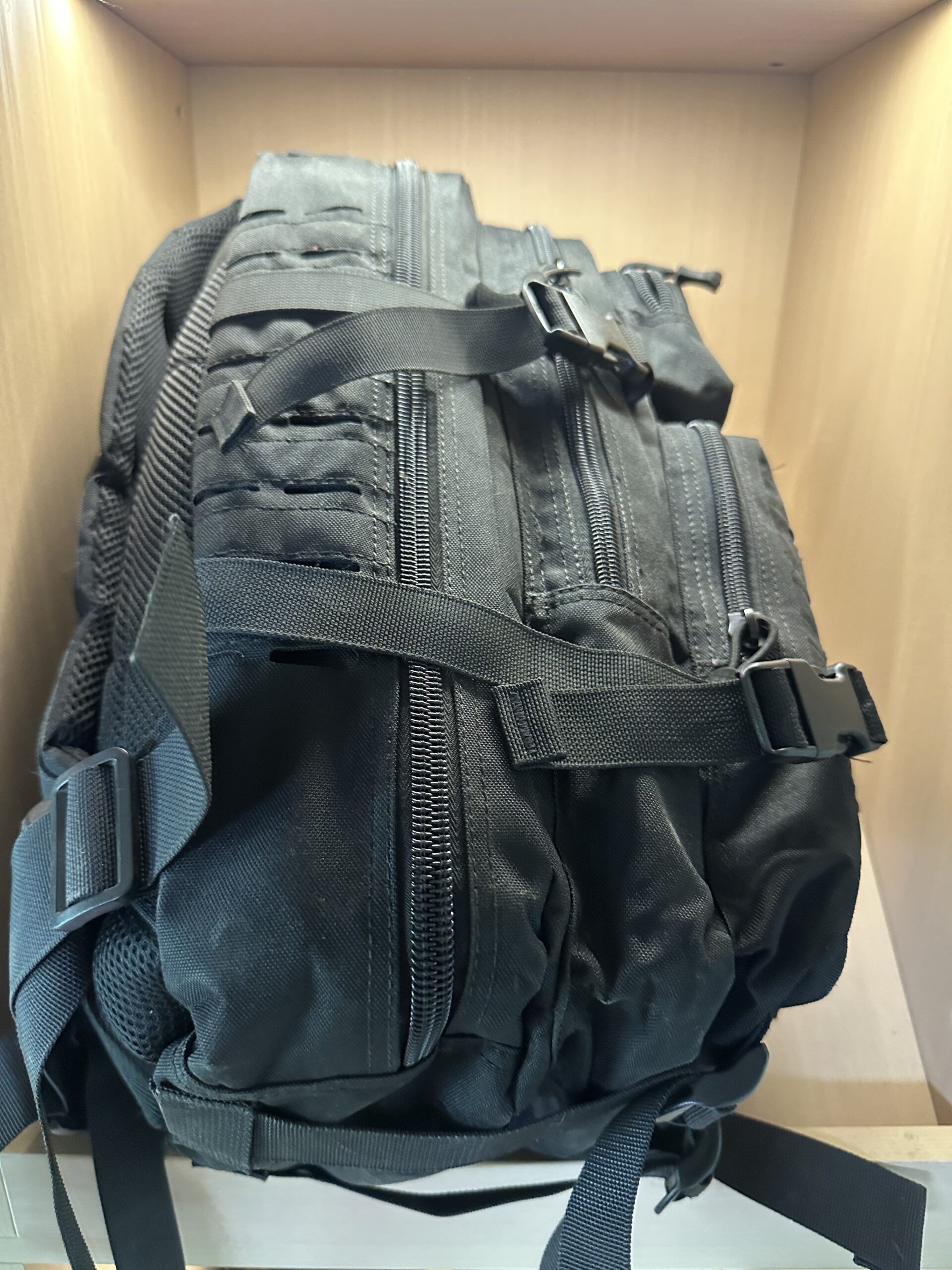 Black tactical MOLLE backpack side view showing zippered compartments, MOLLE webbing, and quick-release buckles