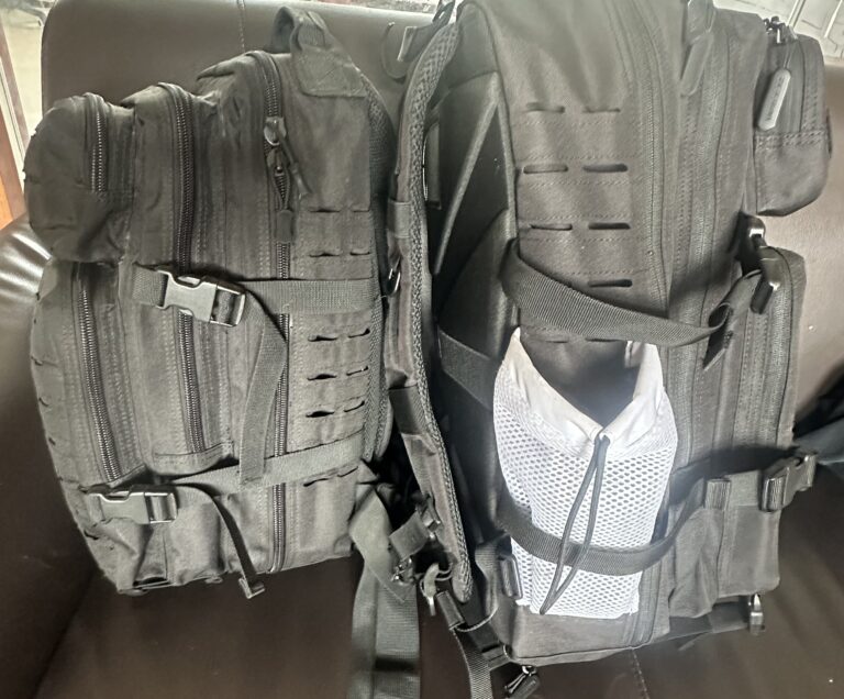 Two black tactical backpacks side by side, one featuring a white mesh side pocket, showing MOLLE webbing and strap details