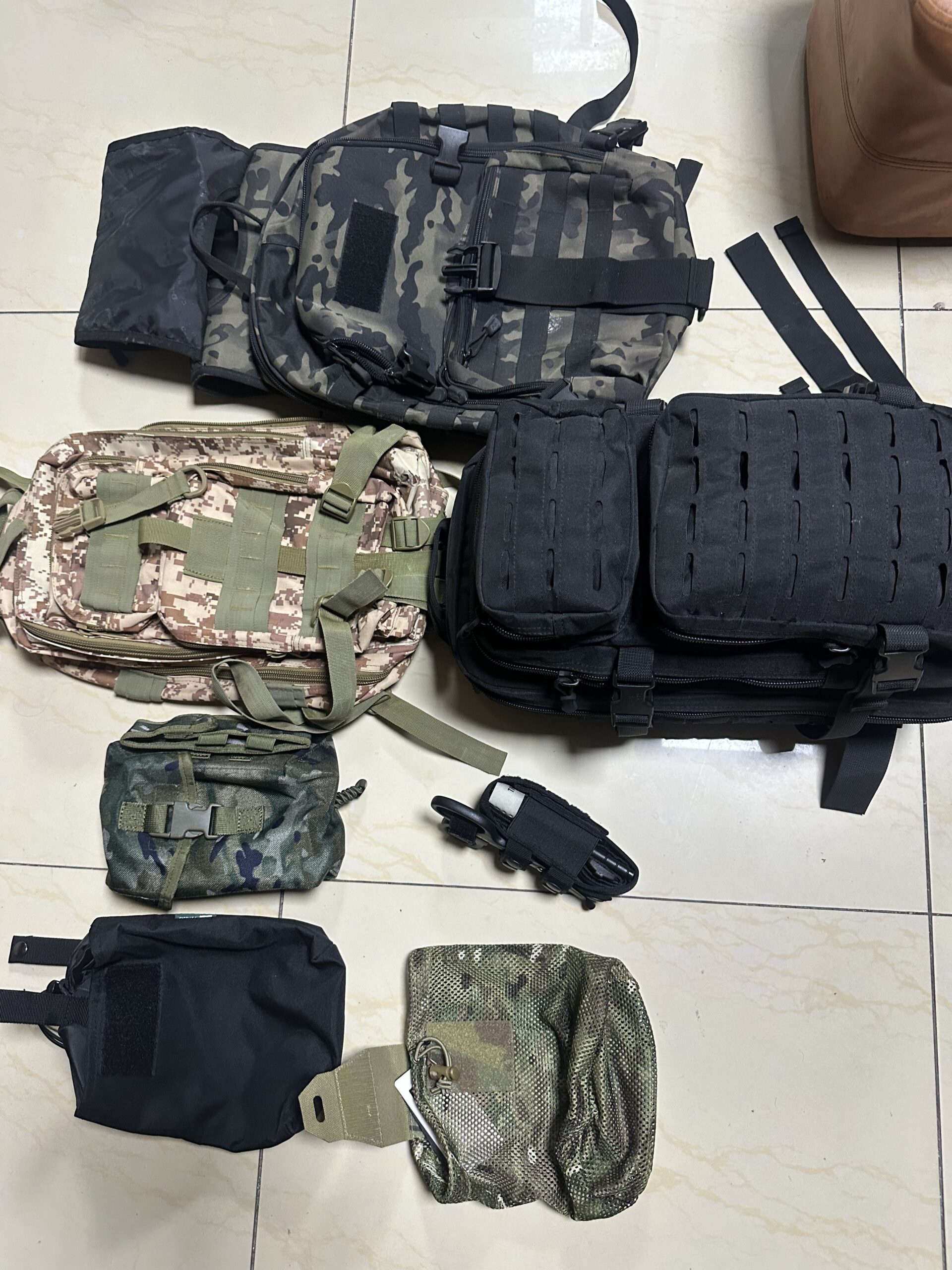 Assortment of tactical gear including a camo backpack, black MOLLE backpack, digital camo backpack, and various utility pouches and holsters laid out for product review